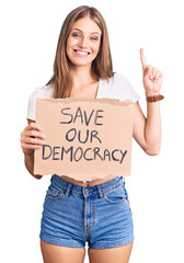 Wall Mural - Young beautiful blonde woman holding save our democracy protest banner surprised with an idea or question pointing finger with happy face, number one