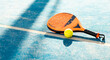 ©  Yistocking - Photo of an orange padel racket and a ball on the ground of a blue outdoor court. Padel concept. Paddle equipment.