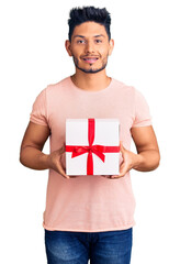 Wall Mural - Handsome latin american young man holding gift looking positive and happy standing and smiling with a confident smile showing teeth