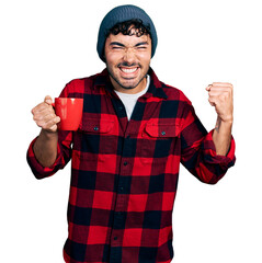 Wall Mural - Hispanic young man with beard drinking a cup of coffee screaming proud, celebrating victory and success very excited with raised arm