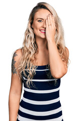 Wall Mural - Young blonde woman wearing casual clothes covering one eye with hand, confident smile on face and surprise emotion.