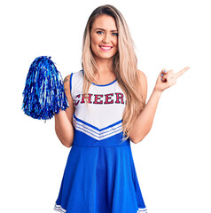 Canvas Print - Young beautiful blonde woman wearing cheerleader uniform holding pompom smiling happy pointing with hand and finger to the side