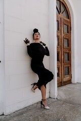 Wall Mural - Stylish woman in the city. Fashion photo of a beautiful model in an elegant black dress posing against the backdrop of a building on a city street