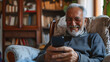 Close-up senior smiling relaxed retired man with beard and glasses sitting comfortably at home on armchair using mobile phone, communication concept