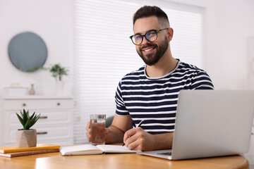 Canvas Print - Young man with glass of water writing in notebook at wooden table indoors