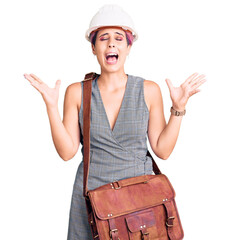 Canvas Print - Young beautiful woman wearing architect hardhat and leather bag celebrating mad and crazy for success with arms raised and closed eyes screaming excited. winner concept