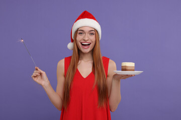 Sticker - Emotional young woman in Santa hat with piece of cake and burning sparkler on purple background