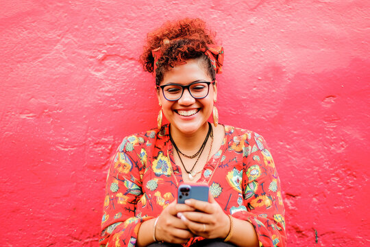 A vibrant portrait of a joyful young woman smiling while texting on her mobile phone against a vivid red background.