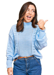 Canvas Print - Young brunette woman wearing casual winter sweater smiling with happy face looking and pointing to the side with thumb up.