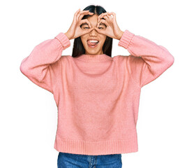 Canvas Print - Beautiful young asian woman wearing casual winter sweater doing ok gesture like binoculars sticking tongue out, eyes looking through fingers. crazy expression.