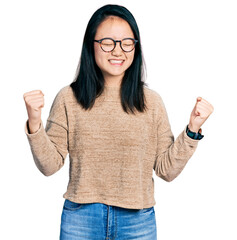 Wall Mural - Young chinese woman wearing casual sweater and glasses very happy and excited doing winner gesture with arms raised, smiling and screaming for success. celebration concept.