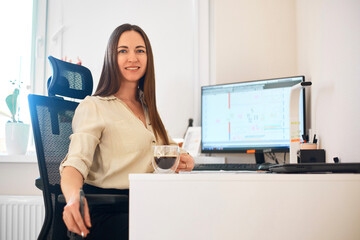 Wall Mural - Portrait of a happy female designer estimator sitting at the table with a notebook and calculator, counting the balance and typing on a laptop, finishing her work with a cup of coffee.