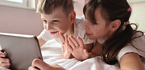 Smiling little children boy and girl lie in bed and look at tablet screen