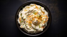  A Bowl Of Mashed Potatoes Topped With Shredded Cheese And Green Onion Sprinkles On A Black Surface.