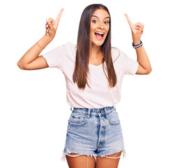 Wall Mural - Young hispanic woman wearing casual white tshirt smiling amazed and surprised and pointing up with fingers and raised arms.