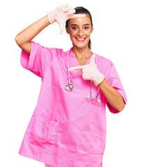 Wall Mural - Young hispanic woman wearing doctor uniform and stethoscope smiling making frame with hands and fingers with happy face. creativity and photography concept.