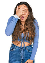 Wall Mural - Young hispanic girl wearing casual clothes peeking in shock covering face and eyes with hand, looking through fingers with embarrassed expression.