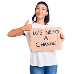 Canvas Print - Young woman holding we need a change banner smiling happy and positive, thumb up doing excellent and approval sign