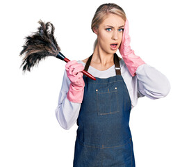 Canvas Print - Beautiful young blonde woman wearing apron holding cleaning duster afraid and shocked, surprise and amazed expression with hands on face