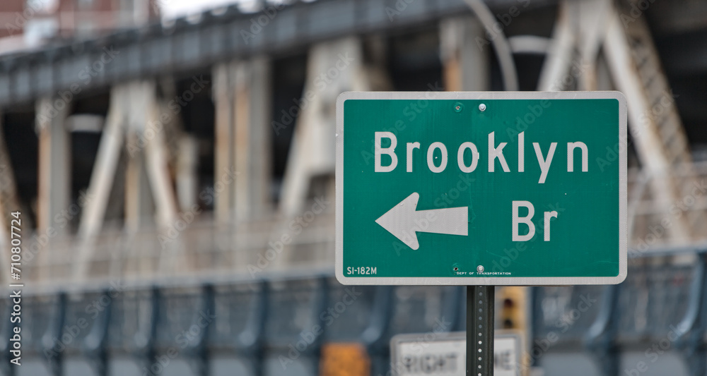 brooklyn bridge sign on the side of the road in downtown brooklyn, new york city (famous landmark travel destination signage in nyc) isolated close up out of focus manhattan bridge background