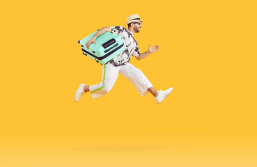 Wall Mural - Full body photo of a funny young happy man in sunglasses carrying his blue suitcase jumping and hurrying up on summer holiday trip on a studio yellow background. Vacation and travel concept.