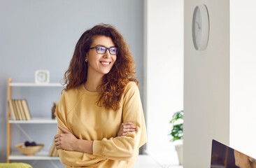 Beautiful female portrait. Happy beautiful long-haired young woman in yellow sweatshirt and glasses standing in a modern room at home, looking away at the window and smiling