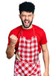 Young arab man with beard wearing cook apron angry and mad raising fist frustrated and furious while shouting with anger. rage and aggressive concept.