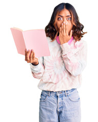 Wall Mural - Young beautiful mixed race woman reading a book wearing headphones covering mouth with hand, shocked and afraid for mistake. surprised expression