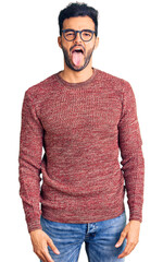 Wall Mural - Young handsome hispanic man wearing winter sweater and glasses sticking tongue out happy with funny expression. emotion concept.