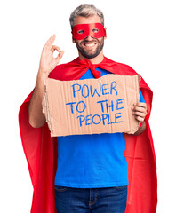 Poster - Young blond man wearing super hero custome holding power to the people cardboard banner doing ok sign with fingers, smiling friendly gesturing excellent symbol