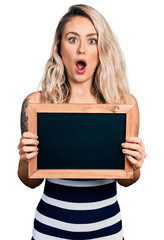 Sticker - Young blonde woman holding blackboard afraid and shocked with surprise and amazed expression, fear and excited face.