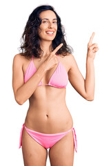 Wall Mural - Young beautiful hispanic woman wearing bikini smiling and looking at the camera pointing with two hands and fingers to the side.