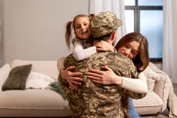 Wall Mural - young soldier of the Ukrainian army in a camouflage uniform returned home to his family, a military dad hugs his little daughter and wife at home