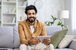 © Liubomir - Portrait of a young Indian man wearing headphones sitting on the couch at home and holding a tablet, smiling and looking at the camera.