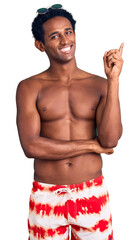 Wall Mural - African handsome man wearing swimsuit and sunglasses with a big smile on face, pointing with hand and finger to the side looking at the camera.