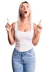 Wall Mural - Young beautiful blonde woman wearing casual sleeveless t-shirt amazed and surprised looking up and pointing with fingers and raised arms.