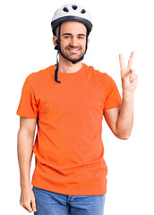 Wall Mural - Young handsome man wearing bike helmet smiling with happy face winking at the camera doing victory sign. number two.