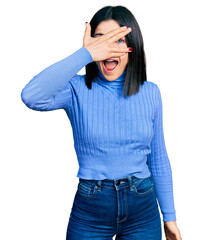 Poster - Young brunette woman with blue eyes wearing casual turtleneck sweater peeking in shock covering face and eyes with hand, looking through fingers afraid