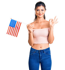 Sticker - Young beautiful woman holding united states flag doing ok sign with fingers, smiling friendly gesturing excellent symbol