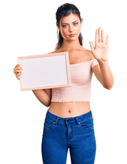 Poster - Young beautiful woman holding empty white chalkboard with open hand doing stop sign with serious and confident expression, defense gesture