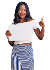 Wall Mural - Young indian girl holding blank empty banner smiling happy and positive, thumb up doing excellent and approval sign