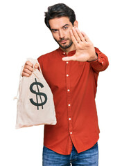 Wall Mural - Young hispanic man holding dollars bag with open hand doing stop sign with serious and confident expression, defense gesture