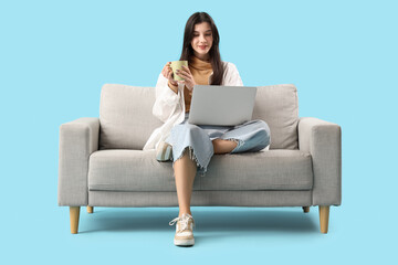 Poster - Beautiful young woman with cup of tea and laptop on sofa against blue background