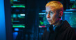 © ihorvsn - Portrait of Caucasian woman hacker with blond short hair sitting in dark room with monitors at desk in front of computer. Turning face to camera and taking on hood. Close up of female hacking system.