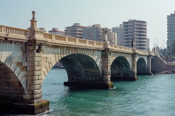 Poster - A picture of a bridge spanning over a body of water with buildings in the background. Suitable for various uses