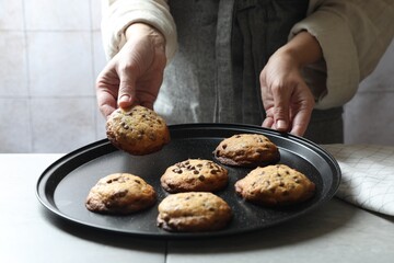 Woman taking delicious chocolate chip cookie from tray at table, closeup