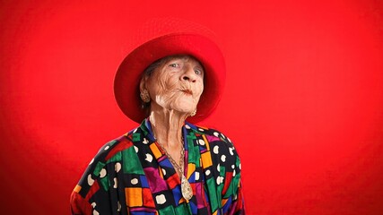 Wall Mural - Funny portrait of mature elderly woman, 80s, having giving OKAY gesture, wearing red hat isolated on red background.