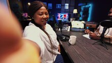 Woman using smartphone selfie camera to film podcast with guest telling interesting story to fans. African american man invited in studio sharing facts, filmed by mobile phone, POV shot