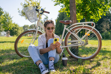 Smiling young hipster woman with bicycle using mobile phone in city park. Talking by video call, using internet