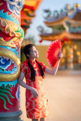 Portrait beautiful cute smiles Asian young woman wearing red traditional Chinese cheongsam decoration and holding a Chinese Fanning for Chinese New Year Festival at Chinese shrine in Thailand